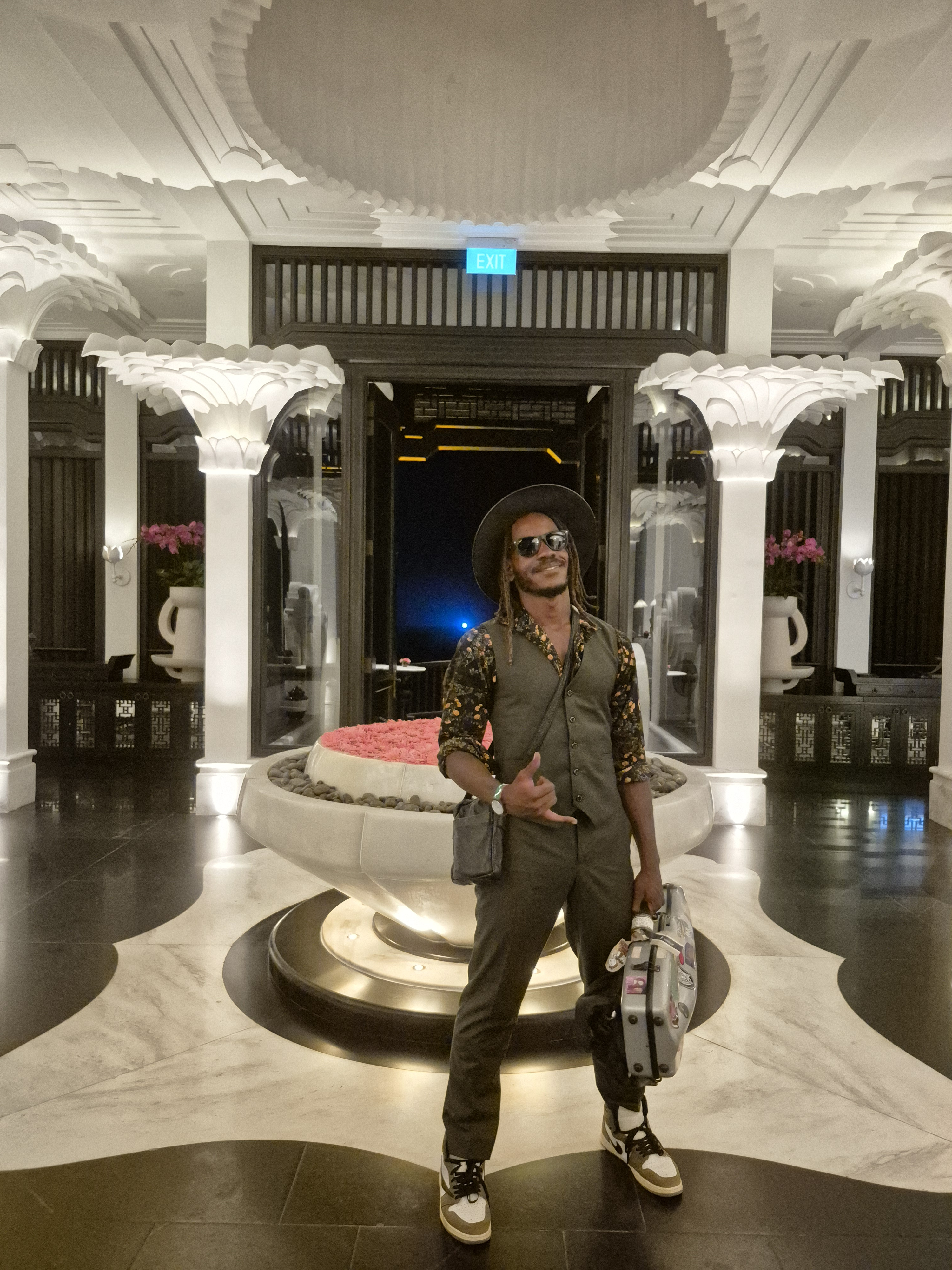 Rahmaan Phillip in a hotel lobby, styled in a vest, brimmed hat and sneakers, holding an instrument case.